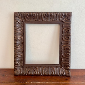 May include: A rectangular dark brown wooden picture frame featuring an ornate, deeply carved relief pattern of scrolling acanthus leaves along the entire border. The frame has a smooth inner edge and a weathered, matte finish on the wood surface.