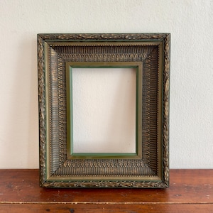 Puede incluir: Un marco de fotos rectangular con un acabado en bronce oscuro y matices verdes. El marco presenta un borde texturizado y ornamentado con tallas en forma de hoja y un borde interior liso de color verde intenso alrededor del centro.