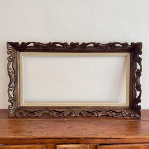 Puede incluir: Un marco de fotos rectangular de madera marrón oscuro con intrincados patrones de volutas talladas a lo largo de los bordes exteriores y un borde interior liso de color crema claro. El marco está vacío, mostrando un centro hueco.