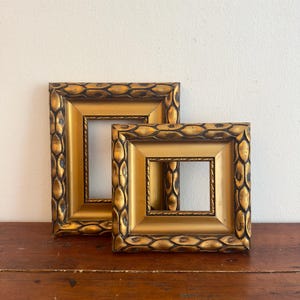 Puede incluir: Dos marcos de fotos rectangulares con un acabado dorado metálico y una pátina oscura y desgastada. Los marcos presentan un patrón repetitivo en relieve similar a hojas en el borde exterior y un borde interior liso y hundido.