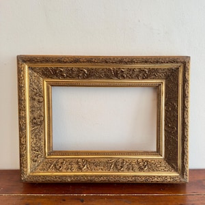 May include: A rectangular picture frame with a metallic gold finish. The frame features an ornate, raised relief pattern consisting of scrolling acanthus leaves, floral motifs, and beaded detailing along the inner and outer edges.