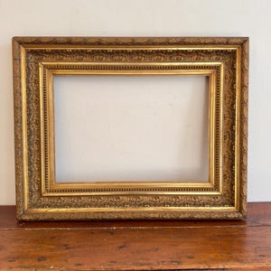 Puede incluir: Un marco rectangular con acabado en oro metálico. El marco presenta intrincadas tallas repetitivas de flores y hojas a lo largo de su borde, complementadas con un detalle de borde interior con cuentas que enmarca la abertura.