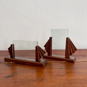 Könnte beinhalten: Zwei dunkelbraune Holzbilderrahmen mit transparenten Glaseinsätzen. Jeder Rahmen hat ein geometrisches Fächerdesign. Die Rahmen werden auf einer Holzoberfläche ausgestellt. Sie sind etwa 15 cm lang.