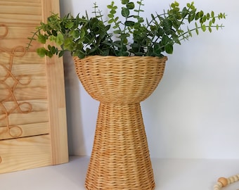 Puede incluir: Una jardinera de mimbre tejido con base de pedestal, llena de tallos de eucalipto verde artificial. La jardinera tiene una parte superior ancha en forma de cuenco y un color natural texturizado. Contraventana decorativa de madera en el fondo.