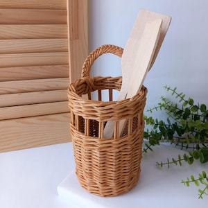 Könnte beinhalten: Ein geflochtener Weidenkorb in hellem Braun mit Griff, der zwei Holzspatel enthält. Der Korb hat eine zylindrische Form mit rechteckigen Aussparungen. Die Spatel sind aus hellem Holz gefertigt.