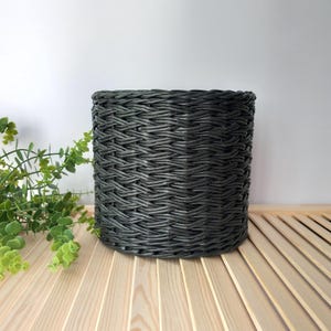 May include: A dark grey woven basket, cylindrical in shape. The basket's tightly woven material creates a textured surface. It rests on a light wooden slatted surface, with green foliage in the background.