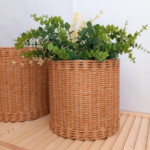 May include: Two woven wicker baskets, one in the foreground and one partially visible in the background. The baskets are filled with green and white leafy plants. The baskets are cylindrical and sit on a light wood surface.