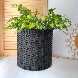 May include: A dark grey woven basket filled with vibrant green leafy branches. The cylindrical basket has a textured, woven design. The arrangement sits on a white surface, with a wooden shutter and decorative sunburst in the background.