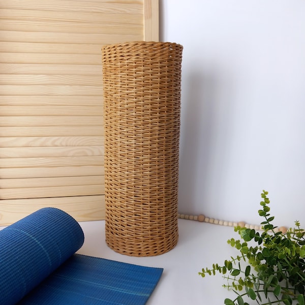Könnte beinhalten: Ein hoher, zylindrischer Korb aus Weidengeflecht in Natur-Beige. Der Korb hat eine gewebte Textur und steht aufrecht. Eine aufgerollte blaue Yogamatte ist teilweise im Vordergrund zu sehen.