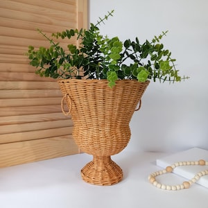 May include: A woven wicker urn-shaped planter with two handles, filled with green leafy faux eucalyptus. The planter is light brown and sits on a white surface. A wooden beaded necklace is in the foreground.