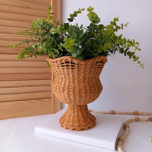 May include: A woven wicker vase with a pedestal base, filled with vibrant green leafy plants. The vase has a flared top and a warm, natural brown colour. The arrangement sits on a white surface, with a wooden shutter in the background.