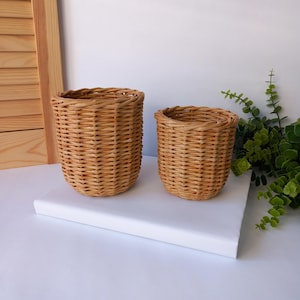Puede incluir: Dos cestas de mimbre tejidas de diferentes tamaños. Las cestas son de color marrón claro y tienen forma redondeada. Están sobre una superficie blanca, con vegetación en el fondo. Estas cestas son probablemente para almacenamiento o decoración.