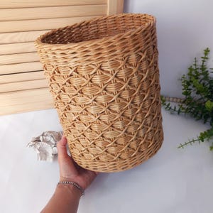 May include: A woven basket with a diamond pattern. The basket is made of light brown wicker and has a cylindrical shape with a slightly angled top. The basket is held by a person's hand, and the background includes a wooden shutter and greenery.