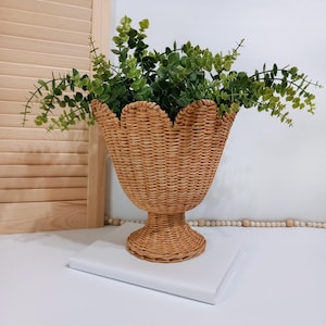 May include: A woven wicker vase with a scalloped rim, filled with green leafy branches. The vase has a pedestal base and sits on a white surface. The natural tones of the vase and greenery create a rustic, decorative accent.