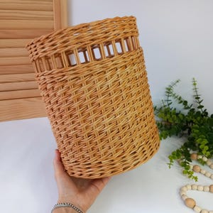 Puede incluir: Una cesta cilíndrica de mimbre tejida a mano, de color marrón natural. La cesta presenta una banda decorativa con recortes rectangulares en la parte superior. La cesta es sostenida por una mano, mostrando su tamaño y textura. Un artículo de almacenamiento decorativo.
