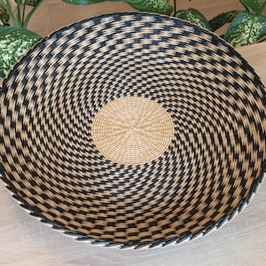 Peut inclure: Un grand panier rond tissé avec un motif en spirale. Le panier présente un centre beige naturel et un motif géométrique noir et beige rayonnant vers l'extérieur. Le bord est fini avec une bordure foncée. Idéal pour la décoration intérieure ou le rangement.