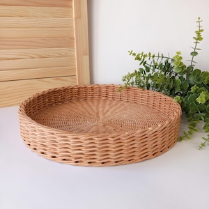 Può includere: Un vassoio rotondo in vimini intrecciato di colore marrone chiaro naturale. Il vassoio ha un design circolare con un motivo intrecciato sulla base e sui lati. Il vassoio ha un diametro di circa 30 cm.