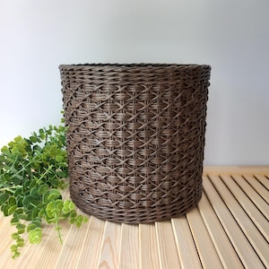 May include: A brown woven basket with a cylindrical shape. The basket has a textured, crisscross pattern and sits on a light-coloured wooden surface. Green leafy plants are visible on the left side of the basket.