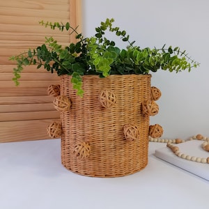 Puede incluir: Una cesta cilíndrica de mimbre, adornada con esferas tejidas, llena de plantas verdes. La cesta, probablemente para decoración, se encuentra sobre una superficie blanca, sugiriendo un elemento decorativo o de almacenamiento.