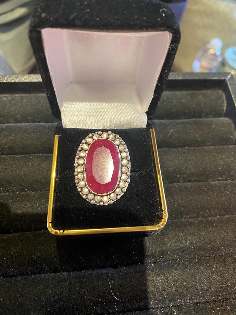 K&ouml;nnte beinhalten: Ein ovaler Ring mit einem gro&szlig;en, tiefroten Edelstein in der Mitte. Der Edelstein ist von einer Bord&uuml;re aus kleinen, wei&szlig;en Steinen umgeben. Der Ring ist in einem silberfarbenen Metall gefasst und wird in einer schwarzen Samt-Schmuckschatulle mit goldenen Akzenten pr&auml;sentiert.