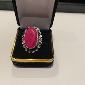 Pode incluir: Um anel oval com uma pedra preciosa rosa vibrante, engastada em metal prateado. O anel &eacute; apresentado numa caixa de joias de veludo preto com acabamentos dourados. O design do anel apresenta uma borda circular detalhada.