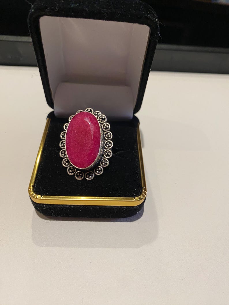 Pode incluir: Um anel oval com uma pedra preciosa rosa vibrante, engastada em metal prateado. O anel &eacute; apresentado numa caixa de joias de veludo preto com acabamentos dourados. O design do anel apresenta uma borda circular detalhada.