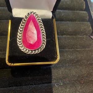 May include: A large, pear-shaped, ruby-colored gemstone ring set in a silver-toned metal with a decorative border. The ring is displayed in a black velvet ring box with gold accents.