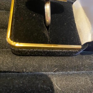 K&ouml;nnte beinhalten: Ein silberner Ring mit einem gro&szlig;en, ovalen, roten Edelstein. Der Edelstein ist in einer dekorativen L&uuml;nette mit kleinen, runden Akzenten gefasst. Der Ring wird in einer schwarzen Schmuckschatulle mit goldenem Rand pr&auml;sentiert.