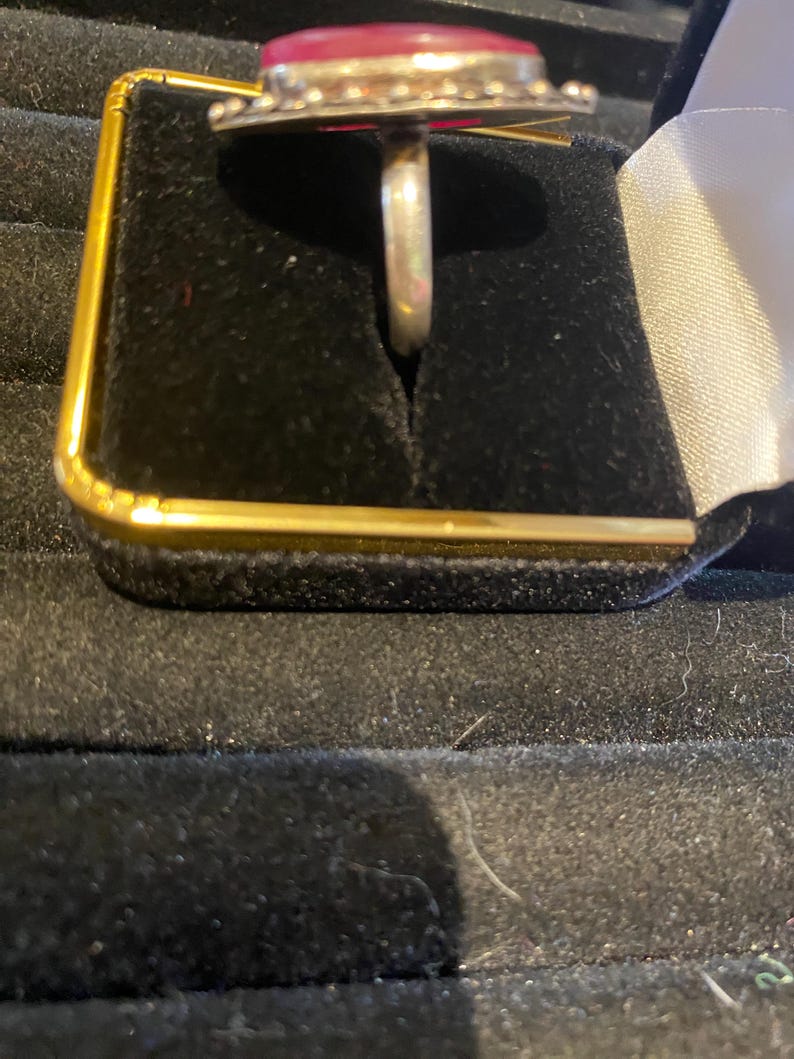 K&ouml;nnte beinhalten: Ein silberner Ring mit einem gro&szlig;en, ovalen, roten Edelstein. Der Edelstein ist in einer dekorativen L&uuml;nette mit kleinen, runden Akzenten gefasst. Der Ring wird in einer schwarzen Schmuckschatulle mit goldenem Rand pr&auml;sentiert.