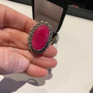 Pode incluir: Anel prateado com uma grande pedra ovalada de cor magenta. A pedra &eacute; rodeada por uma borda decorativa de pequenos detalhes perolados. O anel &eacute; apresentado contra uma caixa de joias preta.