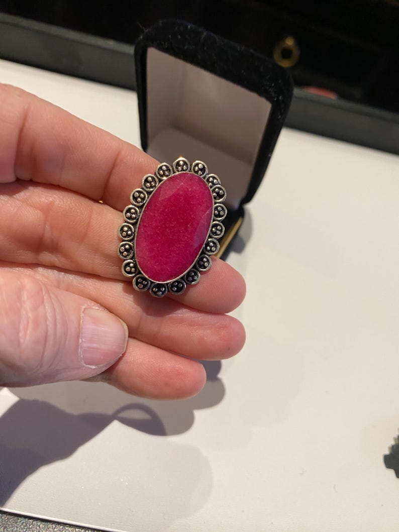 Pode incluir: Anel prateado com uma grande pedra ovalada de cor magenta. A pedra &eacute; rodeada por uma borda decorativa de pequenos detalhes perolados. O anel &eacute; apresentado contra uma caixa de joias preta.