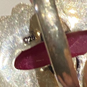 Pode incluir: Close-up de um anel de prata com um design grande, irregular e texturizado. Uma pedra preciosa vermelha escura &eacute; vis&iacute;vel. O anel &eacute; carimbado com "925."