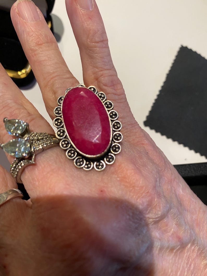 Pode incluir: Um anel grande, em forma oval, com uma vibrante pedra vermelha. A pedra &eacute; engastada em metal prateado com uma borda decorativa de pequenos detalhes redondos. Outro anel com uma pedra verde clara tamb&eacute;m &eacute; vis&iacute;vel.