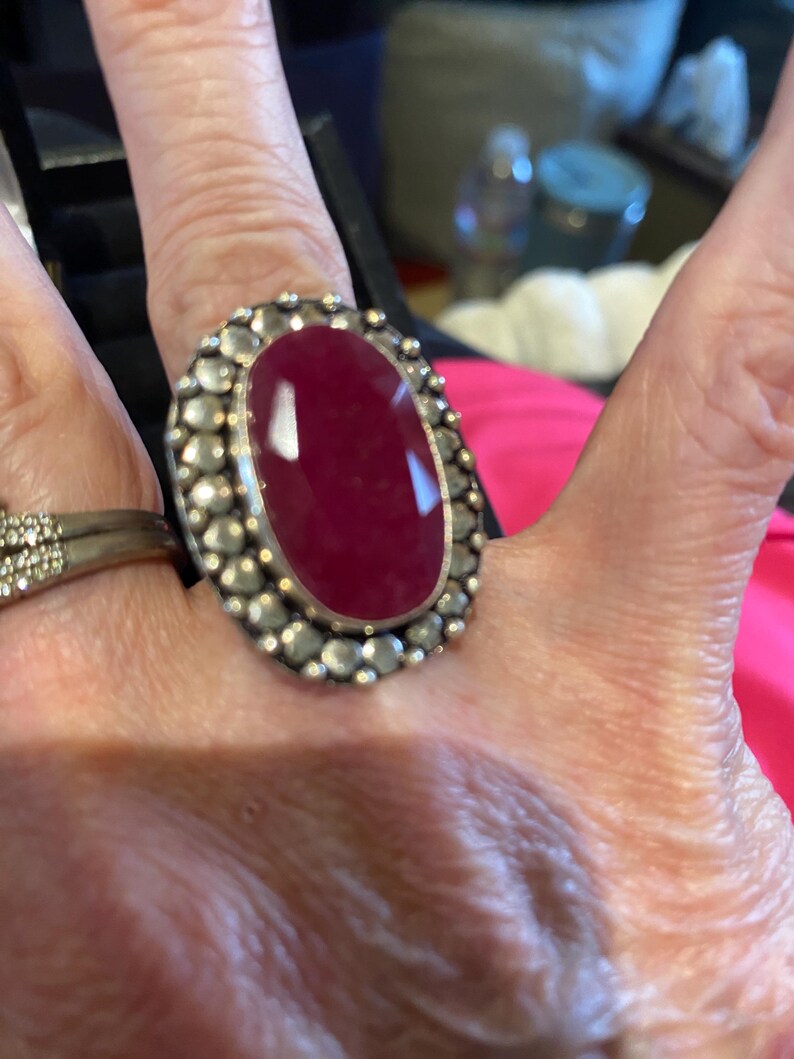 K&ouml;nnte beinhalten: Ein gro&szlig;er, ovaler Ring mit einem tiefroten Edelstein. Der Edelstein ist in einem silberfarbenen Metall gefasst, umgeben von kleinen, runden Verzierungen. Der Ring wird an einem Finger getragen, um sein Design und seine Gr&ouml;&szlig;e hervorzuheben.