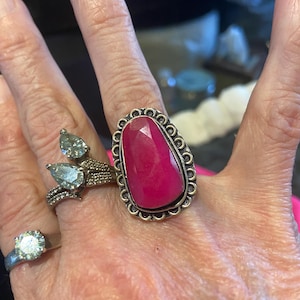 May include: A hand displays several rings, including a large silver ring with a vibrant, oval-cut, ruby-colored stone. Other rings feature clear stones and a double-band design. The rings are set against a neutral background.