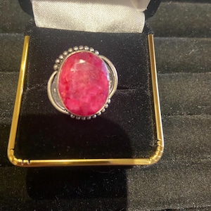 May include: A silver ring with a large, oval, bright pink gemstone. The gemstone is set in a silver bezel with a beaded border. The ring is displayed in a black velvet ring box with gold trim.