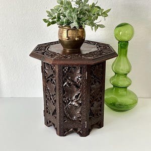 May include: A dark brown octagonal wooden table with intricate carved leaf designs. The table has a small top and folds up for storage. A brass pot with a plant is on top of the table, and a green glass vase is next to it.