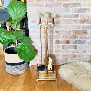 May include: A brass fireplace tool set with a stand. The set includes a poker, shovel, brush, and tongs. The stand has a decorative base and seashell-shaped handles. The tools are arranged in front of a brick wall and hardwood floor.