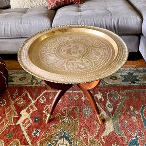 K&ouml;nnte beinhalten: Ein rundes Messingtablett mit aufwendigen, ge&auml;tzten Designs steht auf einem dreibeinigen Holzst&auml;nder mit goldenen Spitzen. Das Tablett verf&uuml;gt &uuml;ber ein zentrales kreisf&ouml;rmiges Design mit chinesischen Schriftzeichen.