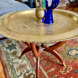 K&ouml;nnte beinhalten: Ein Tabletttisch aus Messing mit einem dekorativen, ge&auml;tzten Design. Der Tisch hat drei Holzbeine mit goldenen Akzenten und steht auf einem gemusterten Teppich.