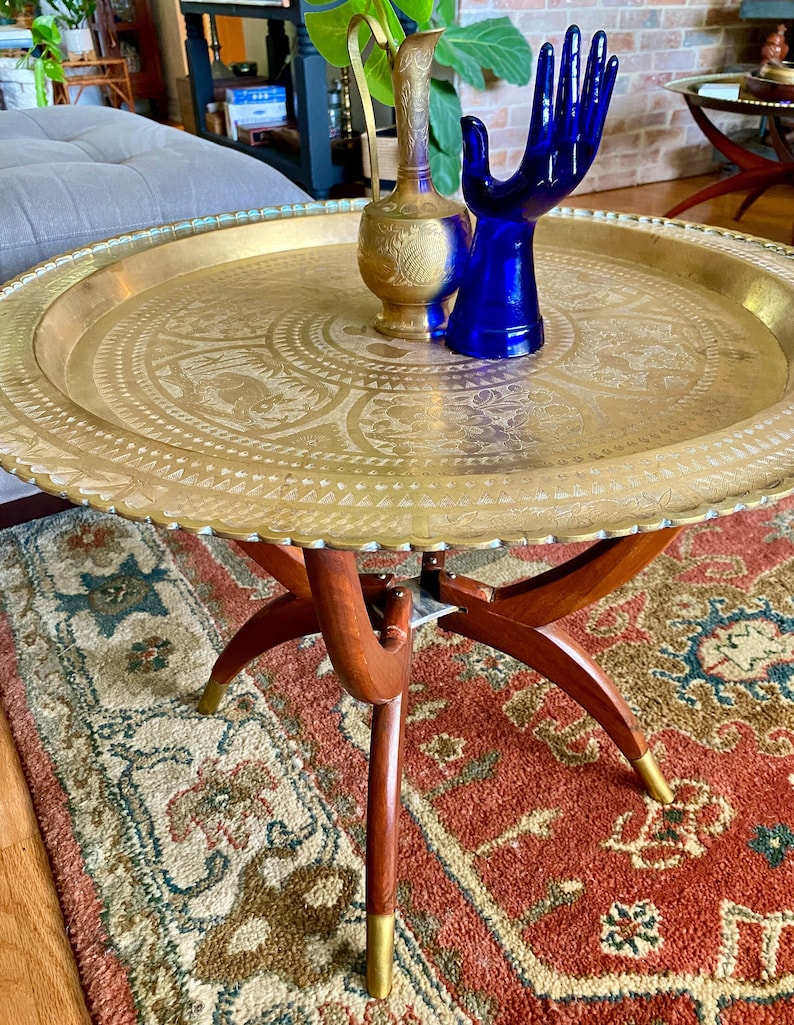 K&ouml;nnte beinhalten: Ein Tabletttisch aus Messing mit einem dekorativen, ge&auml;tzten Design. Der Tisch hat drei Holzbeine mit goldenen Akzenten und steht auf einem gemusterten Teppich.