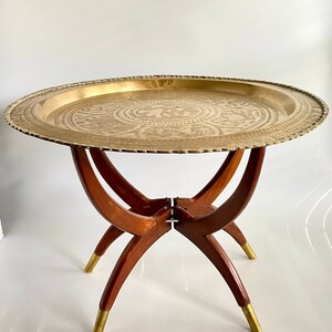 K&ouml;nnte beinhalten: Ein Tabletttisch aus Messing mit einem dekorativen, ge&auml;tzten Design und einem klappbaren Holzgestell. Der Tisch hat eine runde Tischplatte und drei Beine.