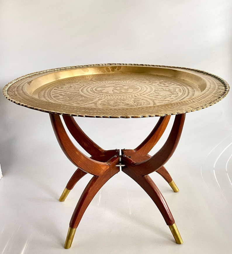 K&ouml;nnte beinhalten: Ein Tabletttisch aus Messing mit einem dekorativen, ge&auml;tzten Design und einem klappbaren Holzgestell. Der Tisch hat eine runde Tischplatte und drei Beine.