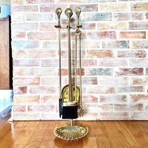 May include: A brass fireplace tool set with a seashell-shaped base. The set includes a poker, shovel, brush, and tongs. The tools have decorative finials and a black-bristled brush. The set is displayed against a brick wall and wooden floor.