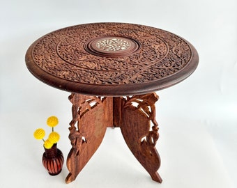 May include: A round, intricately carved wooden table with a floral design. The table has a small, round top with a floral inlay and four legs that are also carved with floral designs. The table is brown in color and is sitting on a white surface.
