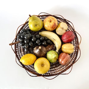 May include: A round, dark brown woven wicker basket filled with a collection of artificial fruit, including a green pear, a yellow banana, a bunch of dark grapes, a lemon, a fig, and several round stone-like fruits in shades of red and yellow.