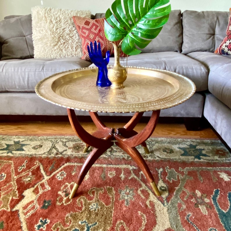 K&ouml;nnte beinhalten: Ein Tabletttisch aus Messing mit einem dekorativen Design und einem Holzgestell. Der Tisch steht auf einem roten und cremefarbenen Teppich mit einer gr&uuml;nen Pflanze in einer goldenen Vase und einer blauen Handskulptur auf dem Tablett.