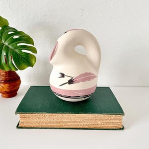 May include: A cream-colored ceramic pitcher with a pink and black feather design. The pitcher has a rounded body with a spout and a handle. The pitcher is sitting on a green book.