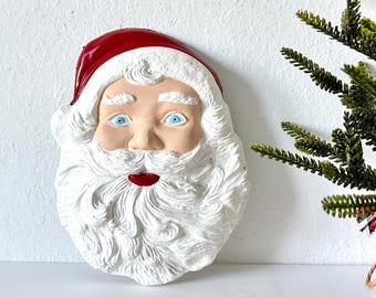 May include: A Santa Claus face ornament with a red hat, white beard, and blue eyes. The ornament is made of a textured material and is likely for Christmas decorations. A small green Christmas tree is in the background.