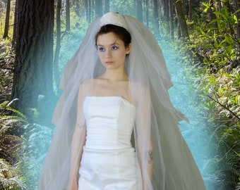 May include: A bride wearing a white strapless wedding dress and a white veil, standing in a forest with a blue and green light effect.
